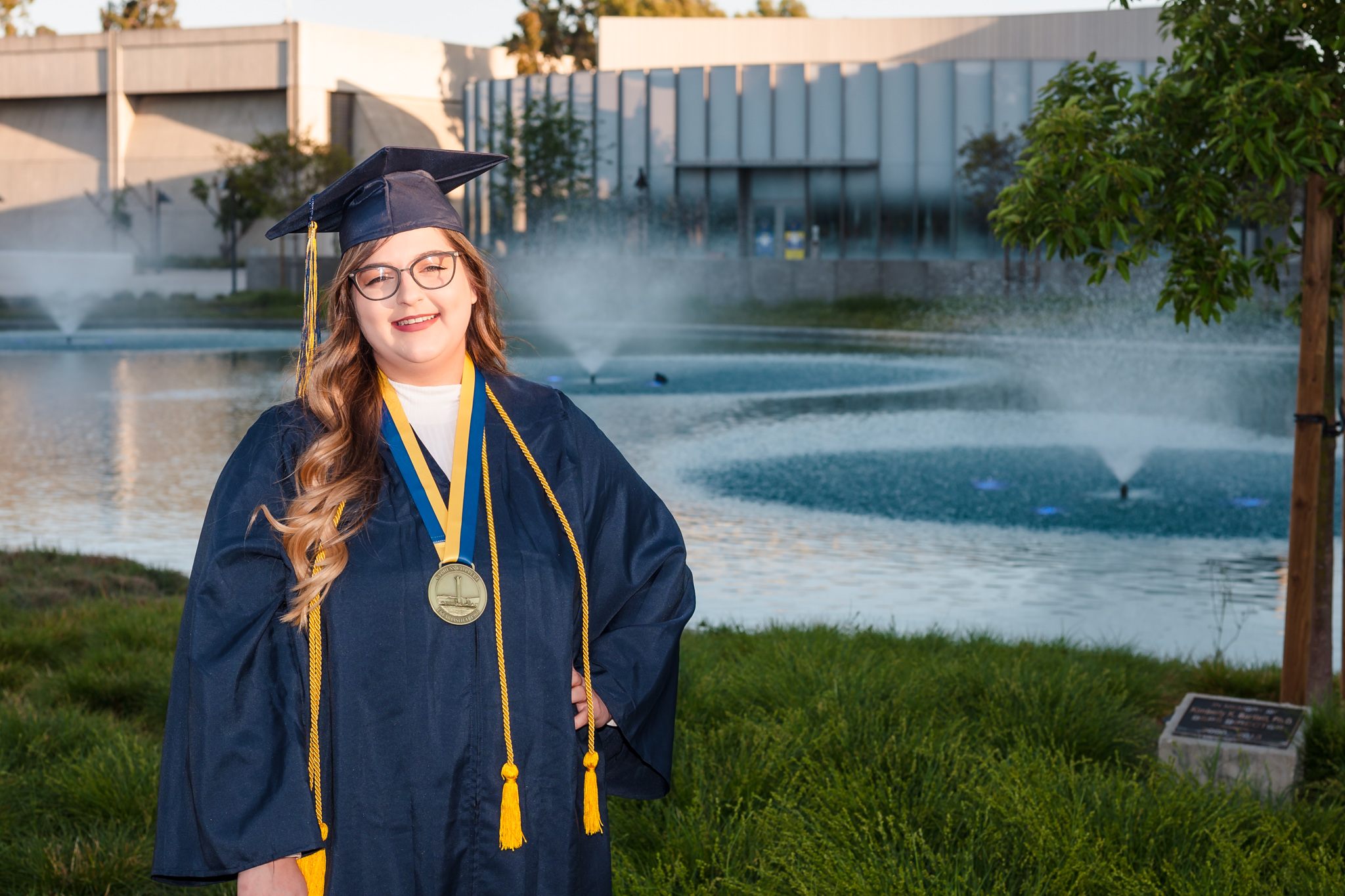 Student Krystal Kosacki wears graduation regalia and poses in front of campus pond.