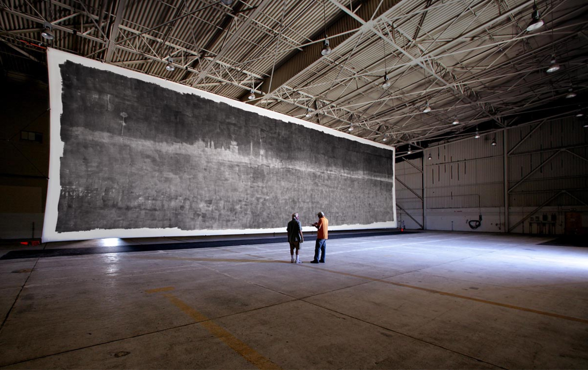 Two adults stand in a large aircraft hangar observing a massive horizontal black and gray abstract photograph suspended from the ceiling.
