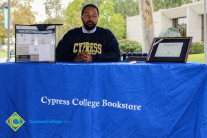 Man wearing a Cypress College sweatshirt sitting at a Cypress College Bookstore table.