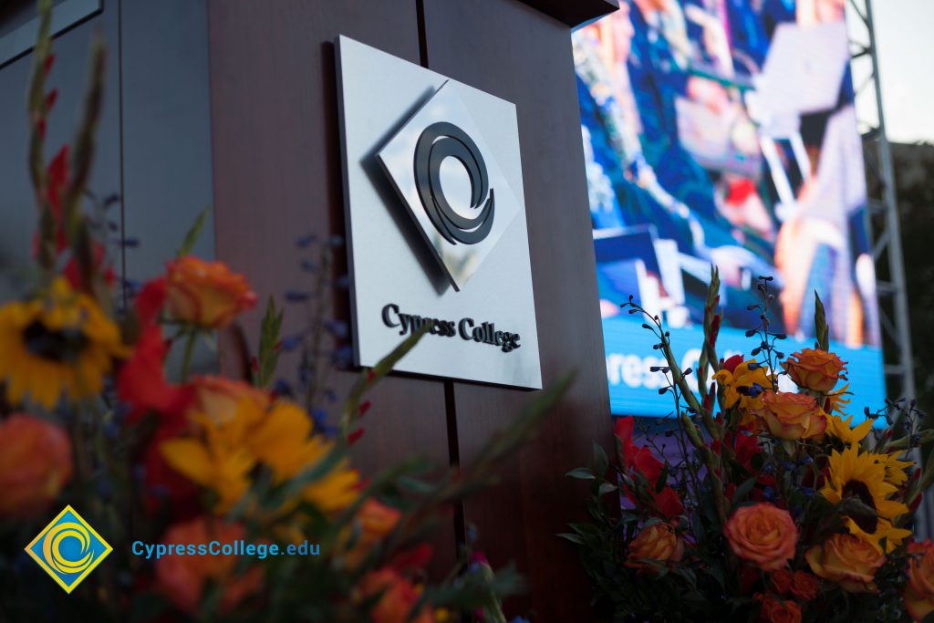 Cypress College sign with logo, big screen monitor, and flowers.