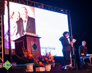 Woman speaking on stage with Holocaust survivors on big screen.