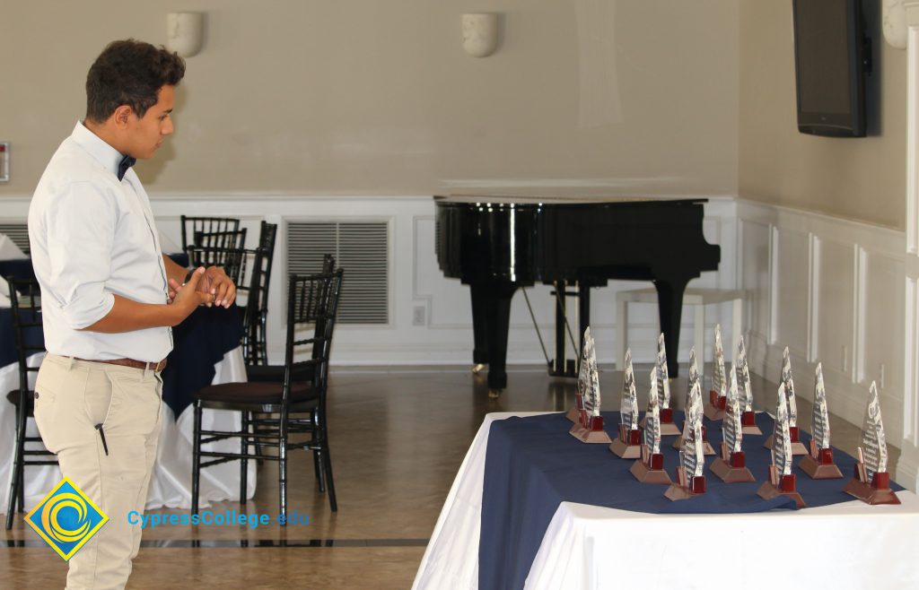 Male student dressed in khaki pants, white long-sleeved shirt rolled up to the elbows, and a bow tie, looking at table of awards