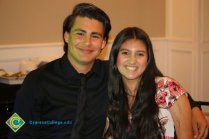 Male student wearing black shirt and tie, with his arm around female student wearing a white and pink floral dress