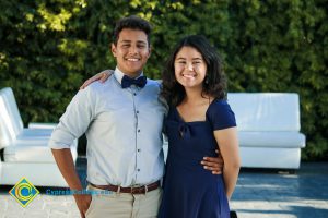 Male student wearing khaki pants, brown belt, light-colored button-down long-sleeved shirt, and navy bow tie, with his arm around a female student who is wearing a navy blue dress.