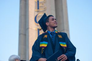 Male wearing graduation cap and gown with blue dress shirt and tie with mult-color sash as he looks to the left