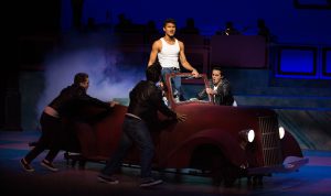 Young man in a white muscle t shirt standing in a car with three young men in leather jackets standing outside car, performing a scene from Grease.