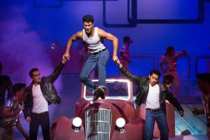 Two young men in black leather jackets helping another young man in a white muscle t shirt jump off hood of car in a scene from Grease.