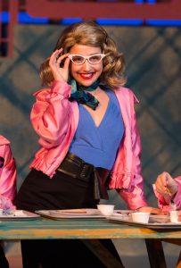 Young lady in pink jacket and white glasses performing in Grease production.