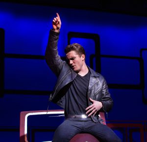 Theater student in black jacket, shirt and pants sitting with right arm pointing up.