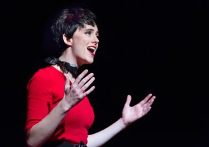 A student with short black hair, red sweater and black scarf, performing in a scene from Grease.