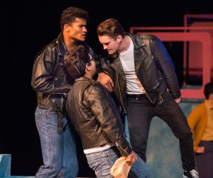 Three young men in leather jackets and jeans performing a scene from Grease.
