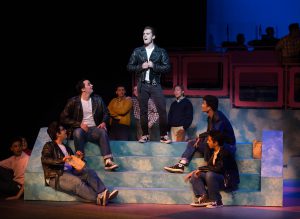 A young man in a black leather jacket and jeans in the spotlight while other actors look on in a scene from Grease.