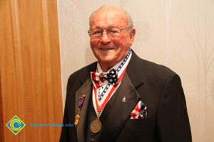Man in glasses with a red, white and blue bowtie and pocket square wearing a medal around his neck and a couple of medals on his lapel.