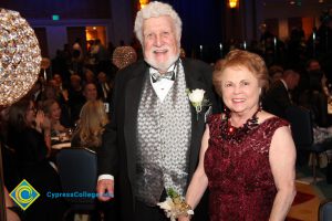 A man with curly white hair and beard in a black suit with grey vest and a woman in a red dress and red hair.