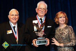 Dr. JoAnna Schilling with two men in suits and tie wearing award medals. One man is holding a Man of the Year award.