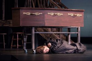 Student on stage, lying on ground wrapped in a blanket underneath a coffin during a performance of Oliver Twist