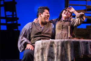 Male and female student sitting at table together, laughing, while performing Oliver Twist