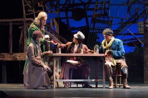 Students on stage, sitting at a table together, in a performance of Oliver Twist