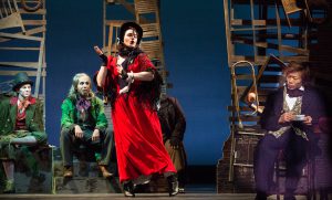 Student in red dress singing on stage with other students looking on while on stage in Oliver Twist