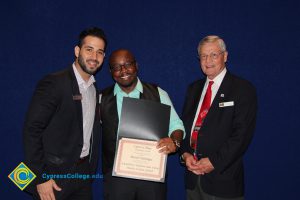 Scholarship recipient in a teal shirt and black vest holding award with a man in a black jacket and dress shirt and a man in a black suit and red tie.