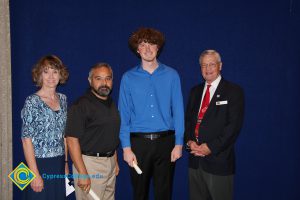 A young man with curly hair and a blue shirt with a man in tan khaki pants, a woman in a blue print shirt and a man in a black suit and red tie.