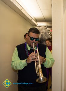 Music professor Gary Gopar dressed in black pants and vest with light green long-sleeved shirt, beads, and sunglasses, playing trumpet while leading Mardi Gras parade