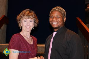 A woman with short brown hair and a red sweater standing with a young man in a black shirt and pink tie.