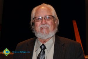 Man with glasses and white hair and beard with brown suit, tan shirt and tie smiling.