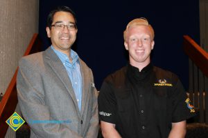 A man in glasses in a grey suit and blue striped shirt and a young man in black shirt and blond hair.