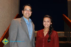 A man with glasses and a grey jacket and blue shirt and a young lady in a red shirt and braided hair.