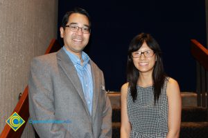 A man in a grey jacket and blue shirt and a young lady with glasses and a black and white dress.