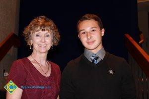 A woman with short brown hair and a red sweater standing with a young man in a black pull over sweater and grey shirt and tie.