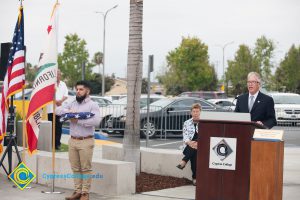 Bob Simpson speaks during 2016 Veteran's Day Anniversary while Pat Ganer watches and a young man holds a folded flag.