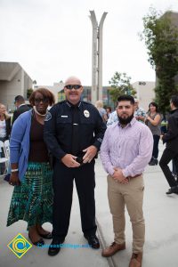 Woman wearing green and black skirt, black shirt, and blue sweater standing next to bald police officer and man wearing khaki pants and buttoned-down shirt.