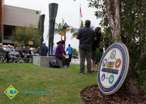 Wall of Remembrance emblem leaning against a tree with people standing and sitting in the background