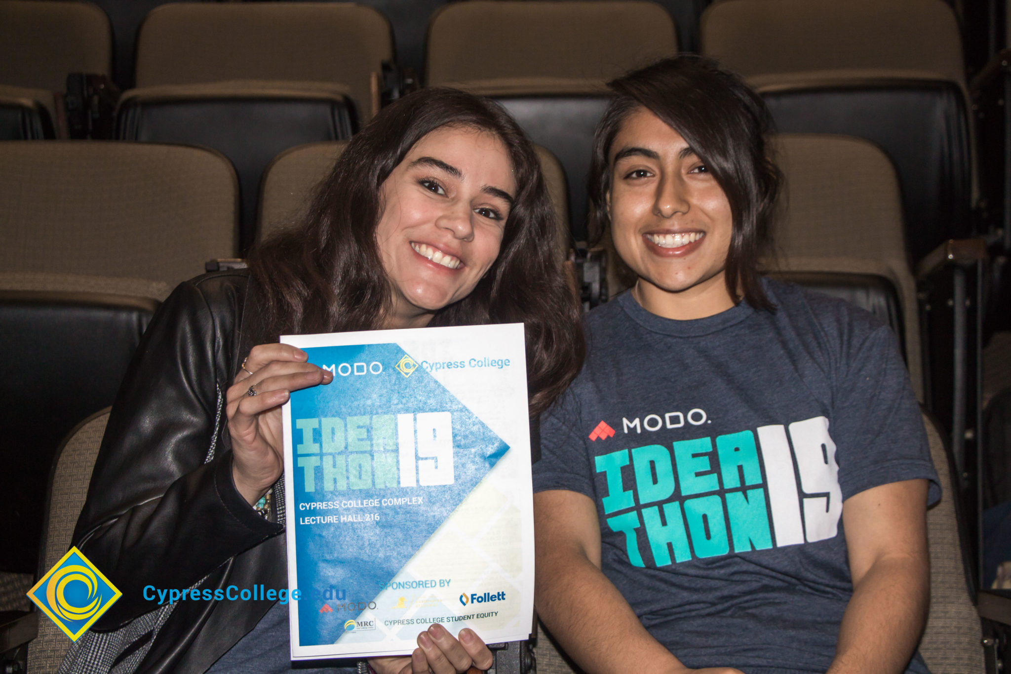 Two young ladies smiling and holding the Modo Ideathon19 flyer.