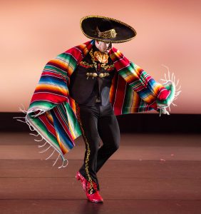 Young man performing folklórico dance, wearing a colorful serape and sombrero.