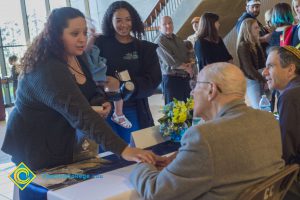 Young lady shakes hands with Holocaust survivor while another young lady holding a toddler and another gentleman look on.