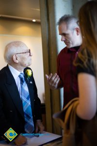 An older gentleman in a black suit, blue tie and yellow boutonnière speaking with a gentleman in a red shirt.