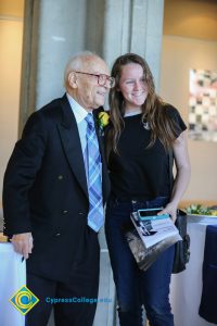 A young lady in long brown hair and a black top standing with a Holocaust survivor in a black suit and blue tie.