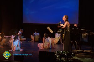 Woman at the microphone as dancers perform at the 2018 Yom HaShoah event.