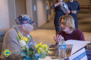 A young man in glasses and red sweater speaking to an older woman at 2018 Yom HaShoah event.