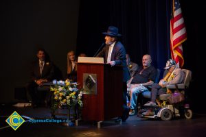 A bearded man in a suit, tie and fedora speaks into a microphone at the 2018 Yom HaShoah event as other speakers look on.