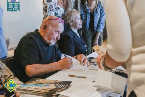 A gentleman with red glasses on his head signs books at the 2018 Yom HaShoah event.