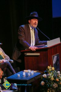 A bearded man in a suit, tie and fedora speaks into a microphone.