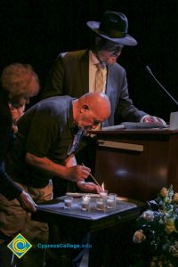 A man lights a candle as another man in a fedora stands at the microphone during the 2018 Yom HaShoah event.