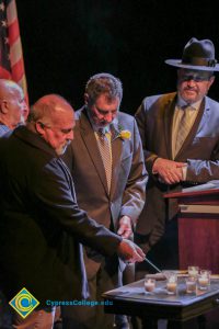 A gentleman with a black coat lighting candles as Clifford Lester and another man in a suit, tie and fedora look on.