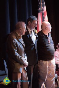 An older gentleman in a brown suit, Clifford Lester and another gentleman in tan khaki pants and a dark pullover standing on stage for the 2018 Yom HaShoah event.