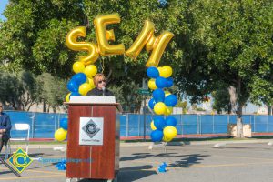 Dr. Schilling at podium with blue and yellow balloon arch and SEM balloons.