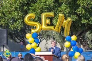 Richard Fee speaking to audience at SEM Groundbreaking ceremony with blue and gold balloons in the background.